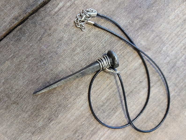 May include: A black cord necklace with a large, silver, nail-shaped pendant. The nail is wrapped with wire near the top.