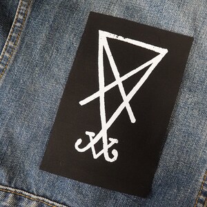 K&ouml;nnte beinhalten: Ein schwarzer rechteckiger Aufn&auml;her mit einem wei&szlig;en, umgekehrten Pentagramm-Symbol. Der Aufn&auml;her ist auf einer blauen Jeansjacke befestigt. Das Symbol hat eine Mittellinie mit sich kreuzenden Linien und gebogenen Elementen unten.