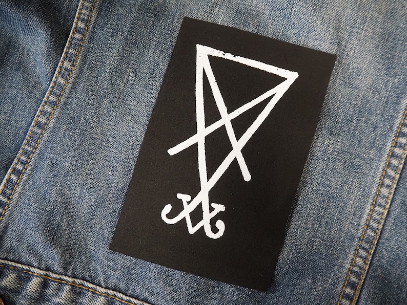 K&ouml;nnte beinhalten: Ein schwarzer rechteckiger Aufn&auml;her mit einem wei&szlig;en, umgekehrten Pentagramm-Symbol. Der Aufn&auml;her ist auf einer blauen Jeansjacke befestigt. Das Symbol hat eine Mittellinie mit sich kreuzenden Linien und gebogenen Elementen unten.