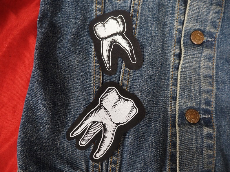 Pu&ograve; includere: Due toppe di denti nere e bianche su una giacca di jeans blu. I denti sono contornati di nero e riempiti di bianco.