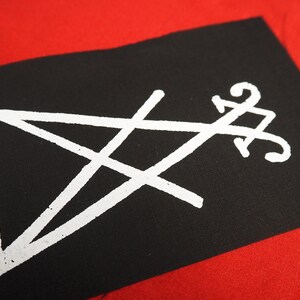 K&ouml;nnte beinhalten: Schwarzer rechteckiger Aufn&auml;her mit einem wei&szlig;en Pentagramm und einem stilisierten Symbol. Der Aufn&auml;her ist vor einem roten Hintergrund platziert, was einen auff&auml;lligen Kontrast erzeugt. Das wei&szlig;e Design hat ein leichtes Used-Aussehen.