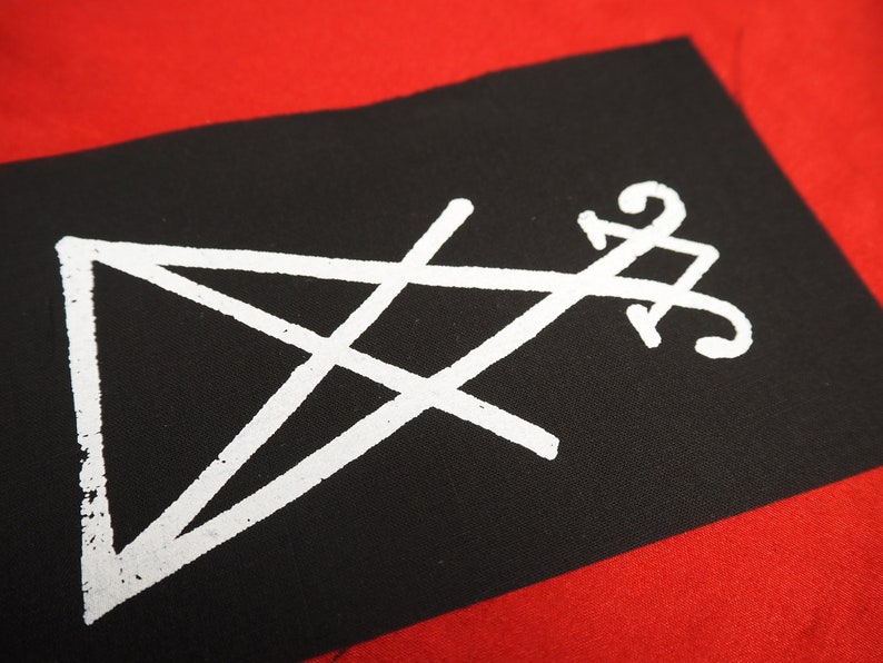K&ouml;nnte beinhalten: Schwarzer rechteckiger Aufn&auml;her mit einem wei&szlig;en Pentagramm und einem stilisierten Symbol. Der Aufn&auml;her ist vor einem roten Hintergrund platziert, was einen auff&auml;lligen Kontrast erzeugt. Das wei&szlig;e Design hat ein leichtes Used-Aussehen.