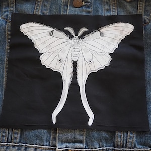 May include: A black fabric patch with a white outline of a moth with long, thin wings. The moth is centered on the patch.