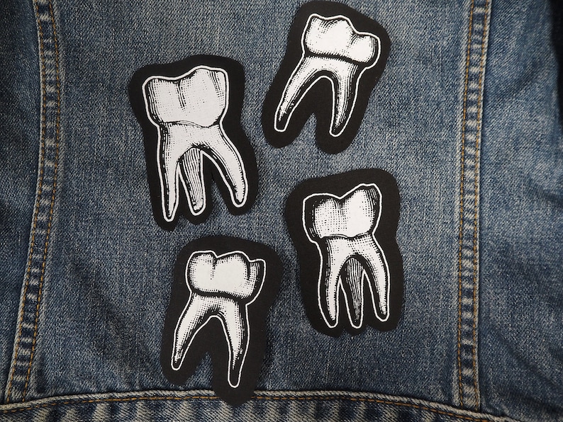 Pu&ograve; includere: Quattro toppe di denti nere e bianche su una giacca di jeans blu. Le toppe hanno la forma di denti e un design dettagliato.