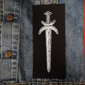 May include: A black patch with a white outline of a sword with a crescent moon on each side.