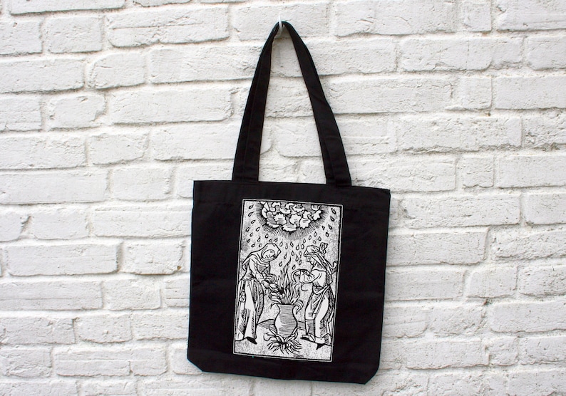 May include: Black tote bag with a white graphic print. The print depicts two robed figures tending to a plant under a cloud with rain. The bag has long black handles and is hanging against a white brick wall.