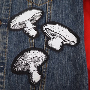 Op de afbeelding: Drie zwarte en witte paddestoel patches op een blauw denim jack. De paddestoelen zijn getekend in een eenvoudige, cartoonachtige stijl.