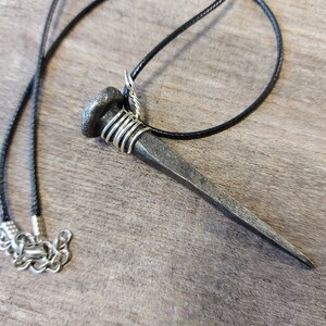 May include: A silver nail pendant necklace with a black cord. The nail is wrapped with silver wire at the top.