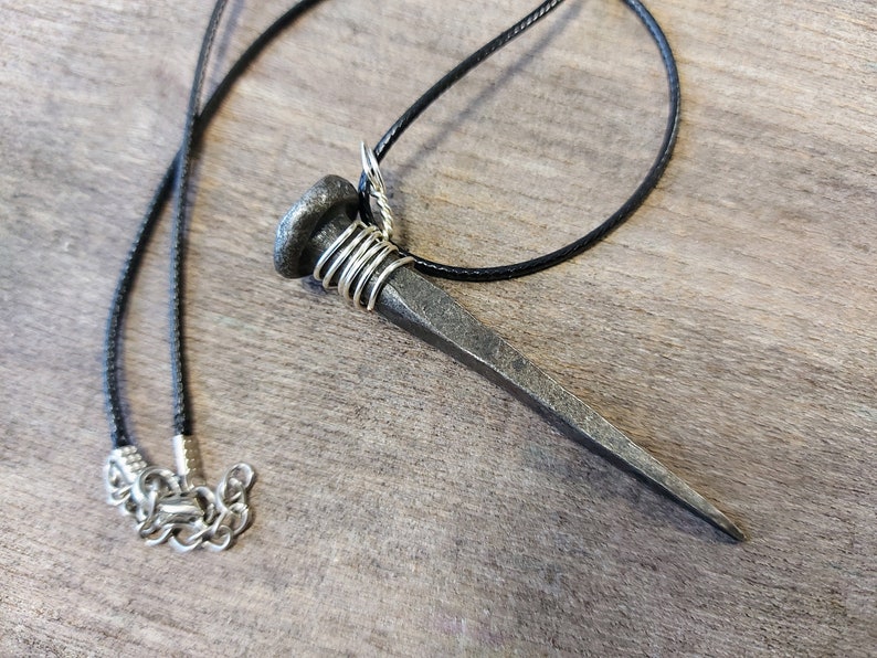 May include: A silver nail pendant necklace with a black cord. The nail is wrapped with silver wire at the top.