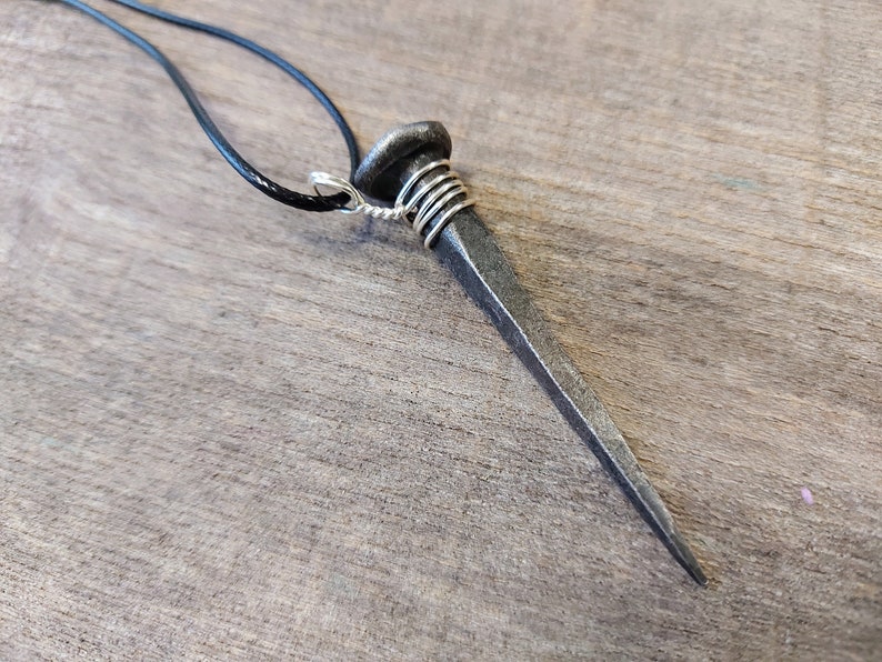 May include: A silver wire wrapped around the head of a large, rusty nail, creating a pendant. The nail is attached to a black leather cord.