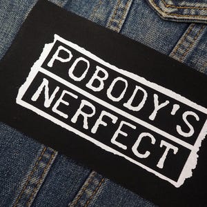 Könnte beinhalten: Ein schwarzer Aufnäher mit den Worten "POBODY'S NERFECT" in weißen Blockbuchstaben. Der Text befindet sich in einem weißen Rahmen im Used-Look. Der Aufnäher befindet sich auf einer Jeansjacke.