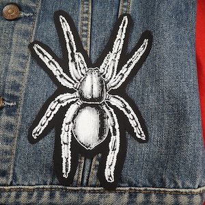 Op de afbeelding: Een zwart-witte spinpatch met gedetailleerde poten en lichaam. De patch is gestreken op een blauw denim jack.