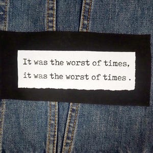 Puede incluir: Chaqueta vaquera azul con un parche negro que muestra la cita "It was the worst of times, it was the worst of times" en fuente de máquina de escribir. Botones metálicos y costuras visibles.