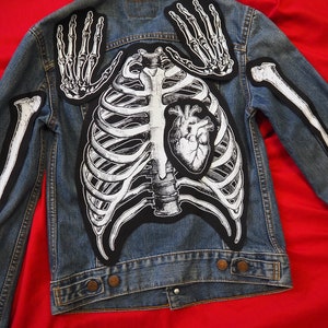 May include: A blue denim jacket decorated with patches showing a human skeleton. The patches feature a rib cage, heart, arm bones, and hand bones, in black and white. The jacket is set against a red backdrop.