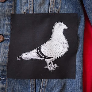 以下が含まれることがあります： デニムジャケット。黒い正方形のパッチには、鳩の白いイラストが描かれています。ジャケットには茶色のボタンと、内側には赤い裏地が見えます。