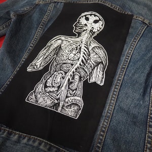 Könnte beinhalten: Eine schwarze Jeansjacke mit einem weißen anatomischen Illustrationspatch auf der Rückseite. Die Illustration zeigt eine menschliche Figur mit inneren Organen und Skelettsystem.