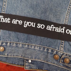 Op de afbeelding: Detailopname van een spijkerjasje met een zwart label. Het label toont de witte tekst "What are you so afraid of?" in een verweerde lettertype. De jas heeft metalen knopen.