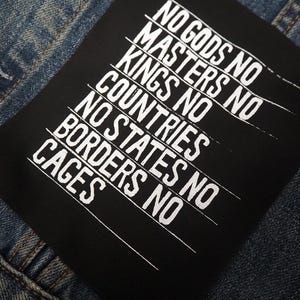 May include: A black patch with white text reading "NO GODS NO MASTERS NO KINGS NO COUNTRIES NO STATES NO BORDERS NO CAGES" is sewn onto a denim jacket. The jacket has a button and visible stitching.