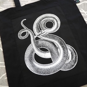 Może przedstawiać: Czarna torba typu tote z szczegółową czarno-białą ilustracją zwiniętego węża. Wzór węża jest wyśrodkowany na przodzie torby, z uchwytami skierowanymi do góry. Torba wykonana jest z wytrzymałej tkaniny.