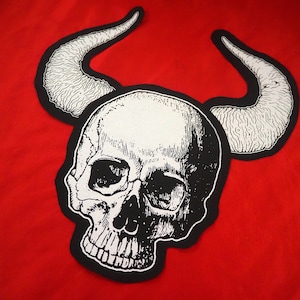 May include: Black and white embroidered patch of a skull with horns. The skull has a detailed design with a textured surface.