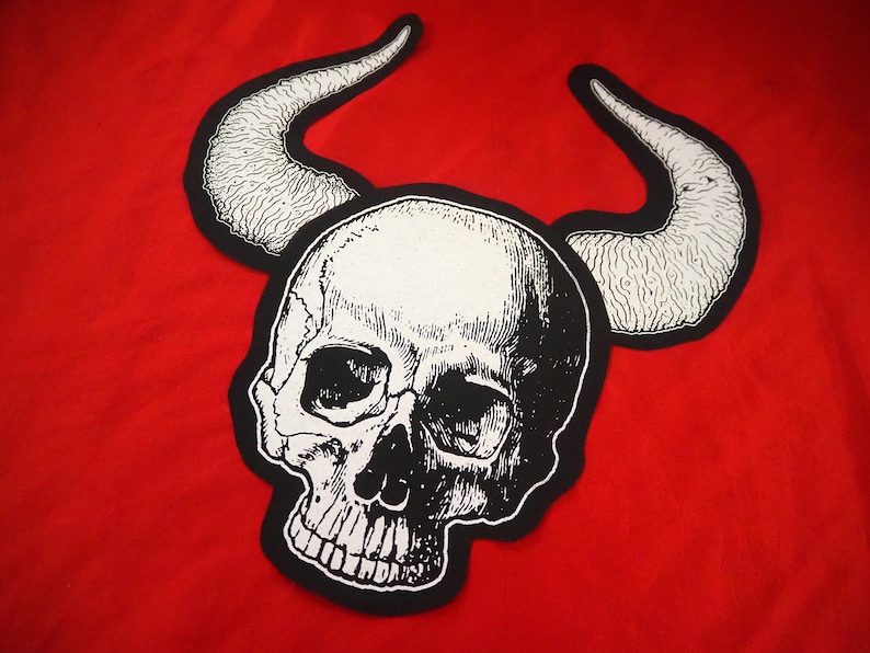 May include: Black and white embroidered patch of a skull with horns. The skull has a detailed design with a textured surface.