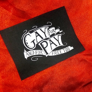 Op de afbeelding: Een zwarte vierkant met de witte tekst "GAY FOR PAY AND FOR FREE TOO" in sierlijke letters. De tekst staat tegen een achtergrond van een feloranje, getextureerde stof.