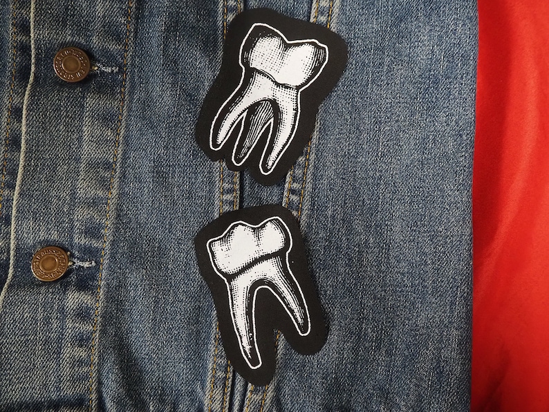 Pu&ograve; includere: Due toppe di denti bianche e nere su una giacca di jeans blu. Le toppe hanno la forma di denti e un design dettagliato.