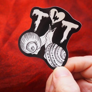 以下が含まれることがあります： 2匹のカタツムリ、ハート、数字の7の白いイラストが描かれた黒いステッカー。カタツムリは互いに向き合い、触角を上げています。ステッカーは赤い背景にあります。
