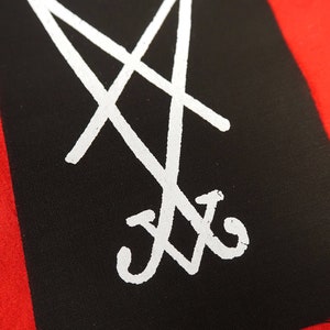 K&ouml;nnte beinhalten: Ein schwarzes Rechteck mit einem wei&szlig;en, stilisierten Symbol vor rotem Hintergrund. Das Symbol ist ein Doppelkreuz mit einem nach unten zeigenden Pfeil und einem gebogenen Boden. Das Bild ist eine Nahaufnahme eines Stoffartikels.