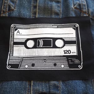 May include: A black and white illustration of a cassette tape on a black fabric patch. The tape is labelled "A." and "120 MIN".
