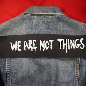 May include: A blue denim jacket with a black patch sewn on the back. The patch has white text that reads "WE ARE NOT THINGS".