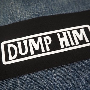 Può includere: Toppa in tessuto nero con testo bianco che recita "DUMP HIM" in un carattere corsivo audace.