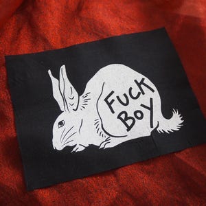 Puede incluir: Un parche rectangular de tela negra con una ilustración serigrafiada en blanco de un conejo de perfil. El conejo tiene orejas largas y bigotes, con el texto "Fuck Boy" impreso en letras negras sobre el cuerpo de la figura del conejo.