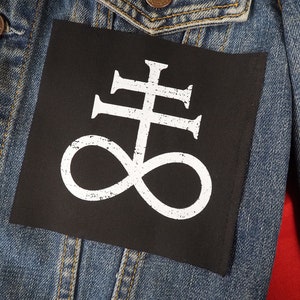 Peut inclure: Un écusson carré noir avec un symbole de croix de Léviathan blanc est cousu sur une veste en jean bleue. L'écusson est centré sur le dos de la veste, et le symbole est une croix stylisée avec un symbole d'infini en dessous.