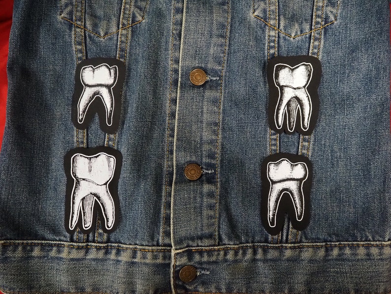 Pu&ograve; includere: Quattro toppe di denti bianchi sono stirate su una giacca di jeans blu. Le toppe sono nere e bianche e presentano un disegno dettagliato di un dente.