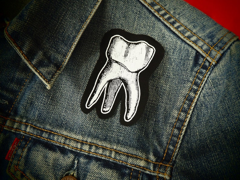 Pu&ograve; includere: Una toppa grafica di un dente bianco su una giacca di jeans blu.