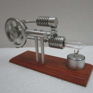 May include: A small, silver, model steam engine with a flywheel and a glass tube. The engine is mounted on a wooden base with a silver burner.