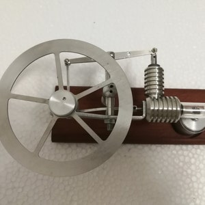 Könnte beinhalten: Ein kleines, silbernes Dampfmaschinenmodell mit einem Schwungrad und einem Glasrohr. Der Motor ist auf einem Holzsockel montiert.