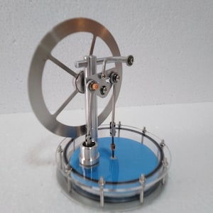 Puede incluir: Un pequeño modelo de motor de vapor de color plateado con una base azul y una cubierta de vidrio transparente. El motor tiene un volante y una biela.