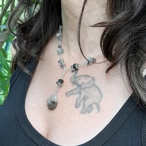 Könnte beinhalten: Eine silberne Halskette mit einem großen, klaren, facettierten Steinanhänger. Die Halskette besteht aus kleinen, grauen und schwarzen Steinen. Die Halskette wird von einer Person mit einem Elefanten-Tattoo auf der Brust getragen.