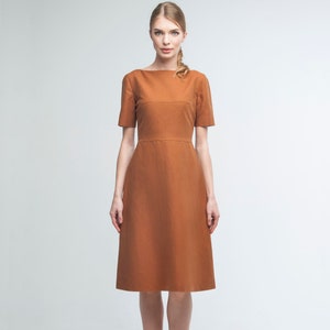 May include: A woman wearing a rust-colored midi dress with a bateau neckline and short sleeves. The dress has a fitted bodice and a slightly flared skirt.