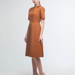 May include: A woman wearing a rust-colored midi dress with a boat neck and short sleeves. She is wearing nude heels.