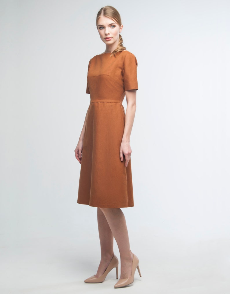 May include: A woman wearing a rust-colored midi dress with a boat neck and short sleeves. She is wearing nude heels.