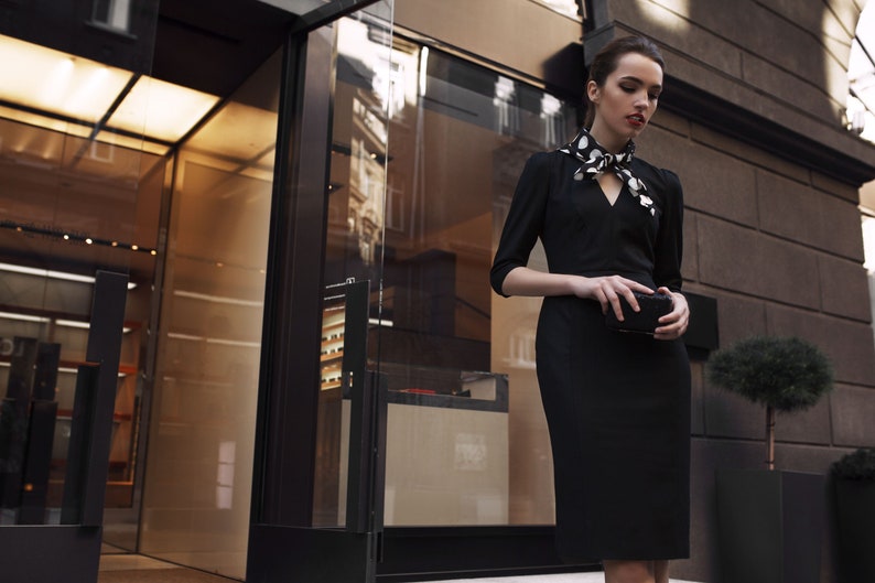 May include: A woman wearing a black dress with a white polka dot scarf and a black clutch.