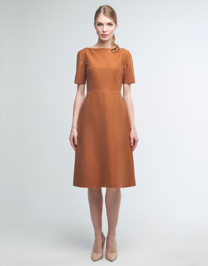 May include: A woman wearing a rust-colored midi dress with a bateau neckline and short sleeves. The dress has a fitted bodice and a slightly flared skirt.