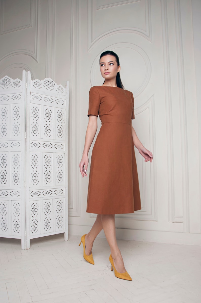 May include: A woman wearing a brown midi dress with a simple design and yellow high heels. She is standing in a room with a white screen behind her.