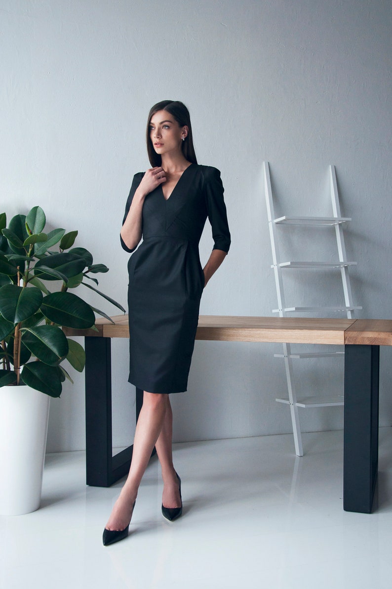 May include: A woman wearing a black knee-length dress with a V-neckline and three-quarter sleeves. She is standing in front of a white wall with a wooden table and a white shelf behind her.