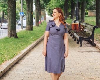 May include: A woman in a gray dress with a ruffled neckline walks on a brick pathway in a park. She is looking to the right of the frame.