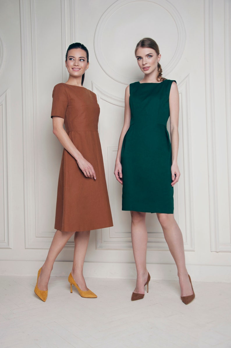 May include: Two women wearing dresses, one in brown and one in green, stand in front of a white wall with decorative molding. Both women are wearing high heels.