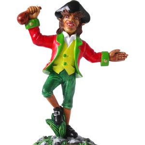 Peut inclure: Une figurine verte, rouge et jaune d'un lutin tenant un pot d'or. La figurine est debout sur une base verte avec le mot "FAITH" écrit dessus.